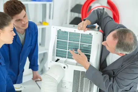 An air conditioner is being repaired by an expert while two apprentices watch and learn.